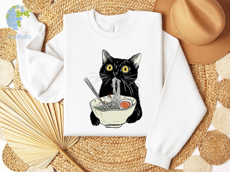 Peut inclure: Sweat-shirt blanc avec un chat noir mangeant des nouilles ramen dans un bol. Le chat a les yeux jaunes et un nez noir.