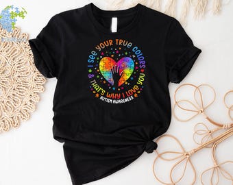 Puede incluir: Camiseta negra con un corazón de pieza de rompecabezas arcoíris y el texto "Veo tus verdaderos colores y por eso te amo Conciencia del autismo".