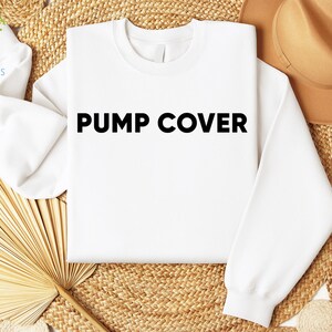Pode incluir: Um moletom branco com o texto "PUMP COVER" em letras pretas em negrito. A pe&ccedil;a &eacute; exibida em uma superf&iacute;cie tecida, acompanhada por um chap&eacute;u marrom e elementos decorativos. A est&eacute;tica geral &eacute; limpa e simples.