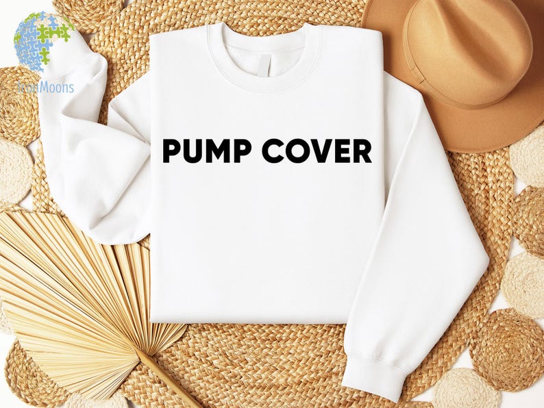 Pode incluir: Um moletom branco com o texto "PUMP COVER" em letras pretas em negrito. A pe&ccedil;a &eacute; exibida em uma superf&iacute;cie tecida, acompanhada por um chap&eacute;u marrom e elementos decorativos. A est&eacute;tica geral &eacute; limpa e simples.