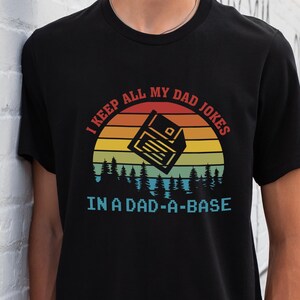 May include: Black t-shirt with a retro sunset graphic and the text "I keep all my dad jokes in a dad-a-base". The graphic features a floppy disk with a sunset behind it.