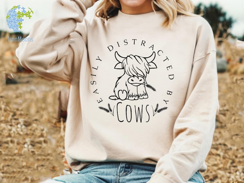 May include: A cream-colored sweatshirt with a black graphic that says "Easily Distracted By Cows" and features a cartoon Highland cow with a floral design.