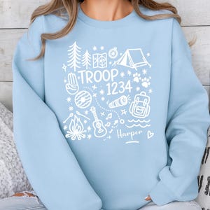 May include: A light blue sweatshirt with a white graphic design featuring camping-themed illustrations. The design includes a tent, trees, a campfire, and the text "TROOP 1234" with a name below. The sweatshirt is a casual style.