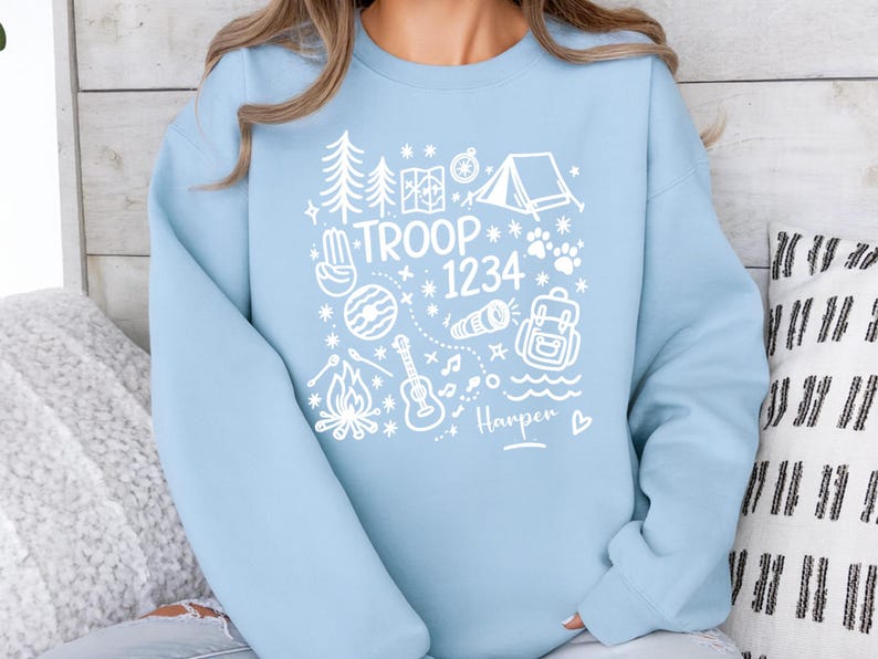 May include: A light blue sweatshirt with a white graphic design featuring camping-themed illustrations. The design includes a tent, trees, a campfire, and the text "TROOP 1234" with a name below. The sweatshirt is a casual style.