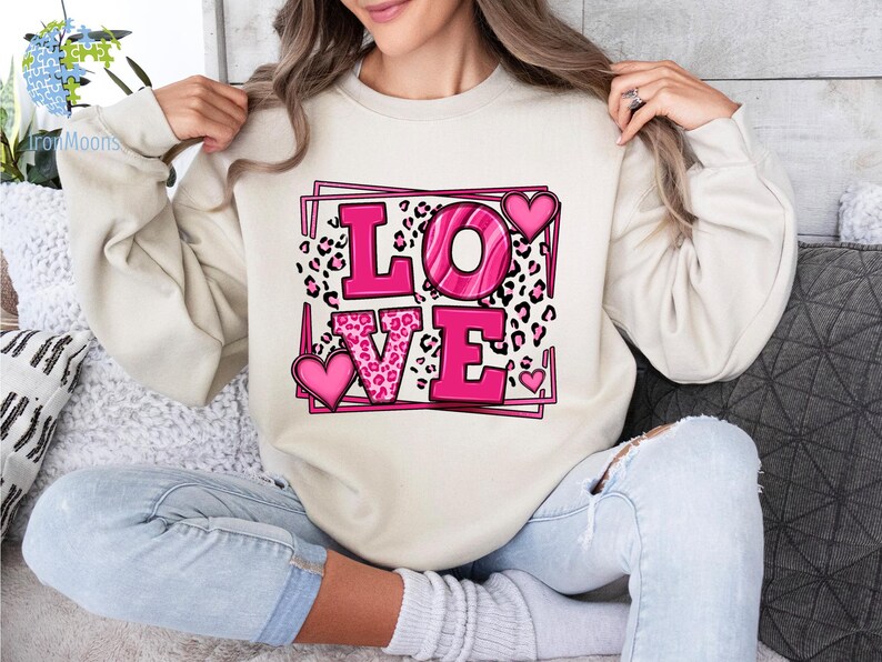 May include: A cream-colored sweatshirt with a pink and black graphic print of the word "LOVE" in a square frame. The letters are outlined in black and have a pink leopard print pattern inside.