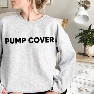 Pode incluir: Um moletom cinza claro com as palavras "PUMP COVER" impressas em letras pretas em negrito no peito. O moletom tem mangas compridas e gola redonda. A imagem mostra uma pessoa usando o moletom.