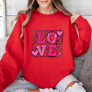 May include: Red sweatshirt with a graphic print of the word "LOVE" in pink letters with a pink leopard print and hearts.