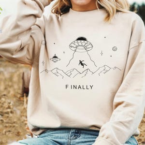 May include: A cream-colored sweatshirt with a graphic of a UFO, a person falling from the sky, and mountains. The text "FINALLY" is printed below the graphic.