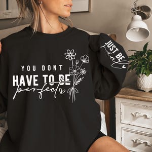 K&ouml;nnte beinhalten: Schwarzer Pullover mit wei&szlig;em Text, der "You don't have to be perfect, just be real" mit einem kleinen Herzen und einem Blumenmuster lautet.
