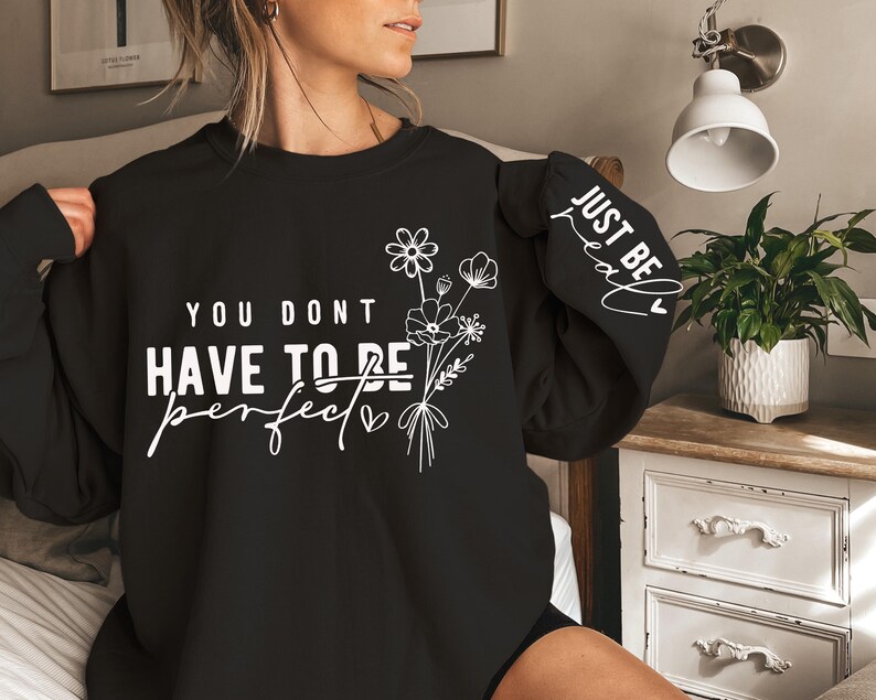 K&ouml;nnte beinhalten: Schwarzer Pullover mit wei&szlig;em Text, der "You don't have to be perfect, just be real" mit einem kleinen Herzen und einem Blumenmuster lautet.