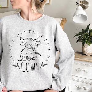 May include: Gray sweatshirt with a black and white illustration of a Highland cow and the text "Easily Distracted By Cows".