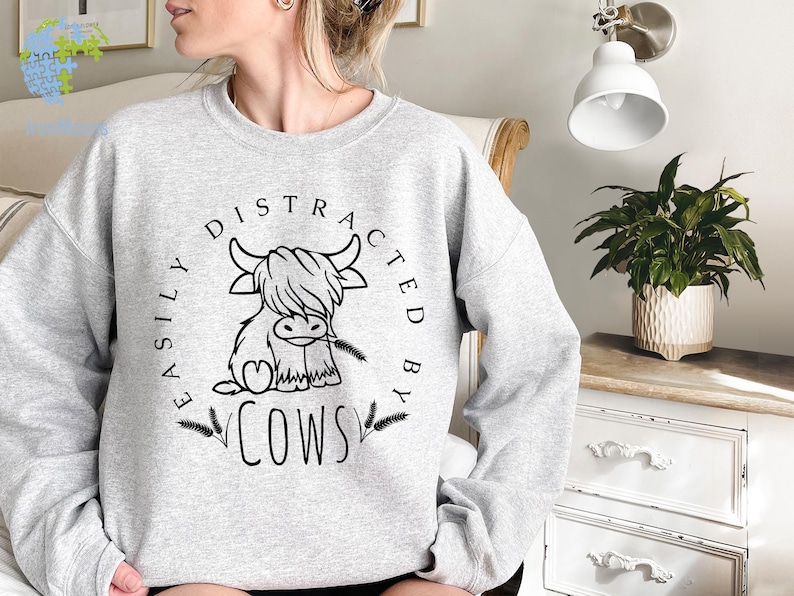 May include: Gray sweatshirt with a black and white illustration of a Highland cow and the text "Easily Distracted By Cows".