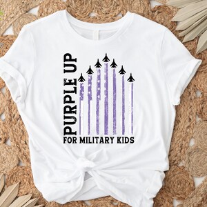 May include: White t-shirt with a purple up for military kids design. The design features a purple and white American flag with black fighter jets flying over it.