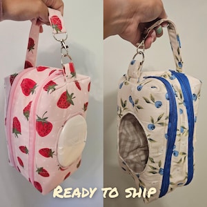 May include: Two rectangular fabric pouches with handles and double zippers. The left pouch is light pink with a strawberry pattern and a white plastic wipe dispenser. The right pouch is cream with a blueberry pattern and a circular opening.
