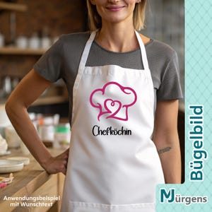 Könnte beinhalten: Weisse Schürze mit einem pinkfarbenen Kochhut- und Herzdesign, mit dem Wort "Chefköchin" in schwarzer Schrift. Die Schürze wird von einer Person in einem grauen T-Shirt getragen.
