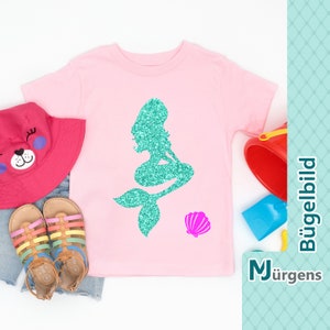 Könnte beinhalten: Pinkes T-Shirt mit einer tealfarbenen Glitzer-Meerjungfrauen-Silhouette und einer pinkfarbenen Glitzer-Muschel. Der Text "Bügelbild" und "M Jürgen" ist rechts im Bild zu sehen.