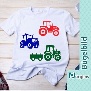 Könnte beinhalten: Ein weißes T-Shirt mit drei Traktor-Silhouetten in Rot, Blau und Grün. Die Traktoren sind alle nach rechts ausgerichtet. Der Text "Bügelbild" und "M Jürgen" ist auf der rechten Seite des Bildes sichtbar.