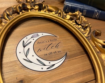 May include: A clear sticker with a crescent moon design, featuring a floral element and the text "there's a little witch in all of us." The sticker is placed on a wooden surface, framed by a gold-colored ornate oval frame. Books are in the background.