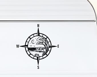 May include: A black vinyl decal applied to a white camper exterior. The design features a compass rose with cardinal directions N, S, E, and W. Inside the circle is a landscape illustration of a small travel trailer, a tree, and a crescent moon.
