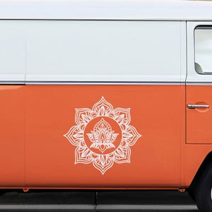May include: A white lotus flower mandala decal on the side of an orange and white vintage van. The mandala is a detailed, symmetrical design. The van has a black tire and a chrome hubcap.