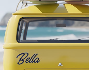 May include: A bright yellow van with the name Bella written in dark blue cursive script on the rear panel. A roof rack holds two surfboards, one light green and one light pink, positioned above the rear window and metal bumper.