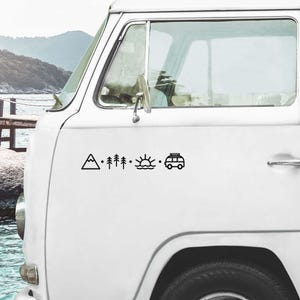 Può includere: Furgone bianco con una grafica nera di una montagna, alberi, sole sull'acqua e un camper. Il furgone è parcheggiato con uno sfondo panoramico di montagne e acqua.
