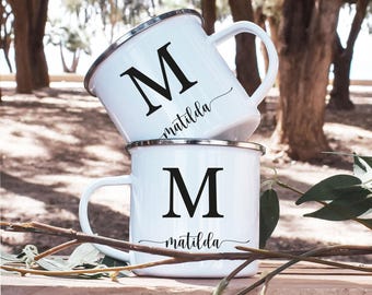 May include: Two white enamel mugs with black lettering. The mugs have a silver rim and a black letter "M" with the name "matilda" written in cursive underneath.