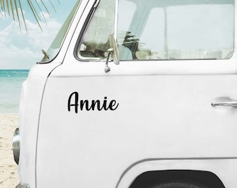 May include: A white van features a black vinyl decal with the name Annie written in a cursive script font on the side panel. The vehicle has a silver side mirror, a silver door handle, and a black tire visible at the bottom.