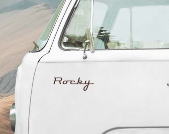 May include: White vintage van with the word "Rocky" in a brown script font on the side. The van has a side mirror and a window. The background shows a mountain range.