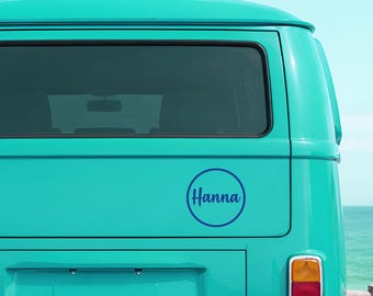 May include: A circular dark blue vinyl decal featuring the name Hanna in a cursive script font, applied to the rear panel of a bright turquoise van. The sticker design consists of a simple dark blue outline circle enclosing the text.