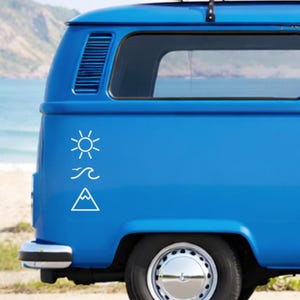 Könnte beinhalten: Ein blauer Van mit einer weißen Grafik von Sonne, Wellen und einem Berg. Der Van parkt an einem Strand, mit dem Meer und den Bergen im Hintergrund. Das Rad und die Radkappe des Vans sind sichtbar.