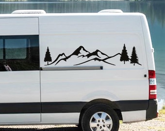 May include: Black mountain and pine tree decal applied to the side of a white van. The decal features a silhouette of a mountain range with two pine trees on either side. The van is parked near a lake.