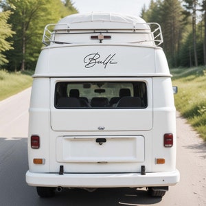 May include: A white vintage camper van, seen from behind, driving on a road. The word "Bulli" is written in cursive on the back. The van has a roof rack and a pop-up top. The background shows trees and a road.