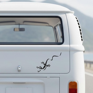 Könnte beinhalten: Ein schwarzer Eidechsenaufkleber auf der Rückseite eines weißen Vans. Das Eidechsendesign hat einen langen, geschwungenen Schwanz. Der Van hat eine große Heckscheibe und eine Seitenlüftung.