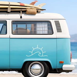 Puede incluir: Furgoneta vintage azul claro y blanca con tablas de surf en el techo. La furgoneta tiene una calcomanía blanca de sol y ola. La escena sugiere un tema de playa, con el océano de fondo.
