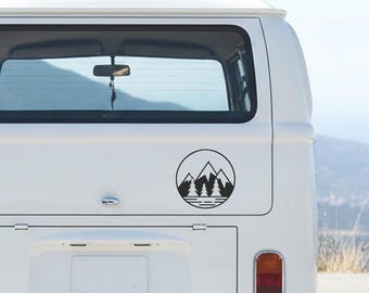May include: A black and white circular sticker featuring a mountain range, trees, and a body of water. The sticker is affixed to the rear of a white van. The design is simple and graphic, suitable for outdoor enthusiasts.
