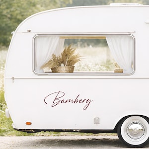 May include: A cream-coloured vintage caravan with the name "Bamberg" in burgundy script. The caravan has a window with sheer white curtains and a basket of dried plants inside. The wheel has a white hubcap.