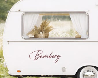 May include: A white vintage-style travel trailer featuring a rectangular window with white curtains and a decorative plant inside. The exterior side of the trailer displays the word Bamberg in a dark red cursive script font.