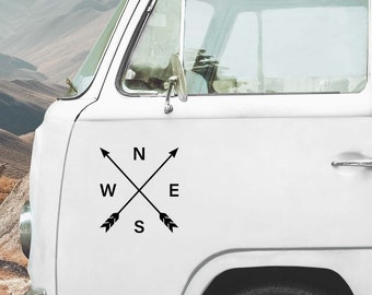 May include: A white van features a black decal on its side panel. The decal displays two crossed arrows forming an X shape, with the letters N, S, E, and W positioned between the arrow shafts to represent the four cardinal compass directions.