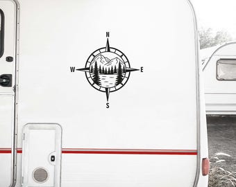 May include: A black vinyl decal applied to the side of a white camper trailer. The decal features a circular compass design with cardinal directions N, S, E, and W, enclosing a graphic illustration of a mountain range, pine trees, and a lake.