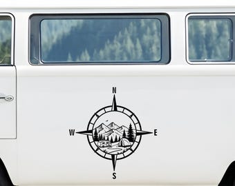May include: A black vinyl decal applied to the side of a white van. The design features a compass rose with cardinal directions N, S, E, and W. Inside the circle is a landscape illustration of mountains, pine trees, a small camper, and a tent.