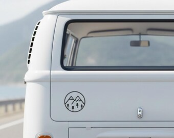 May include: A black circular sticker featuring a mountain and tree design. The sticker is affixed to the back of a white van. The design includes two mountain peaks and three trees inside a circle.