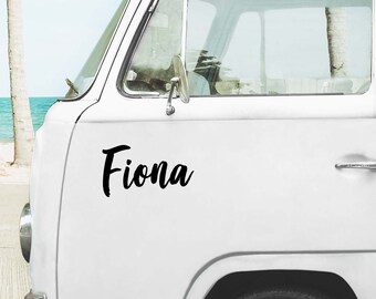 May include: A white van featuring a black vinyl decal on the side panel. The decal displays the name Fiona in a bold, cursive script font. The vehicle has a large front window, a side mirror, and a visible wheel well, with a coastal scene visible.