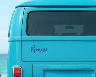 May include: A turquoise vintage van with the name "Bonnie" in a cursive font on the back. The van has a rectangular rear window and a red and orange tail light. The background shows a glimpse of the ocean and sky.