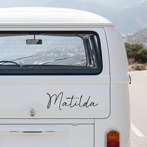 Puede incluir: Parte trasera de un vehículo blanco con el nombre "Matilda" en escritura cursiva negra. El vehículo tiene una ventana trasera, una luz de matrícula y un parachoques. El fondo muestra una carretera y colinas.