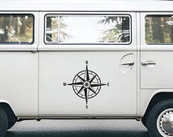 May include: A white van featuring a black compass rose decal on the side panel. The compass design includes a central star shape with cardinal direction letters N, S, E, and W positioned at the top, bottom, right, and left points respectively.