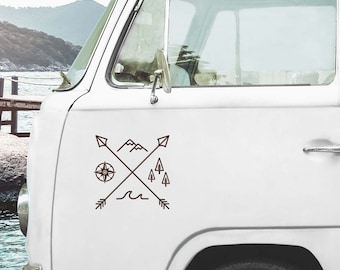 May include: A brown decal featuring a compass, mountains, trees, waves, and crossed arrows, applied to the side of a white van. The design evokes a sense of adventure and travel, with a minimalist, outdoorsy aesthetic.