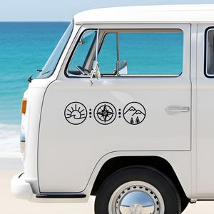 May include: White van with three black circular decals. The decals depict a sunrise over waves, a compass, and a mountain scene with trees. The van is parked near a body of water.