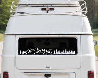 May include: A white vinyl decal applied to the rear window of a white van. The decal features a detailed landscape design with jagged mountain peaks on the left side and a dense forest of pine trees on the right, with a small bird silhouette.