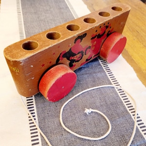 May include: A wooden toy train with red wheels and a string attached. The train has six compartments and a colorful illustration of two figures on the side.